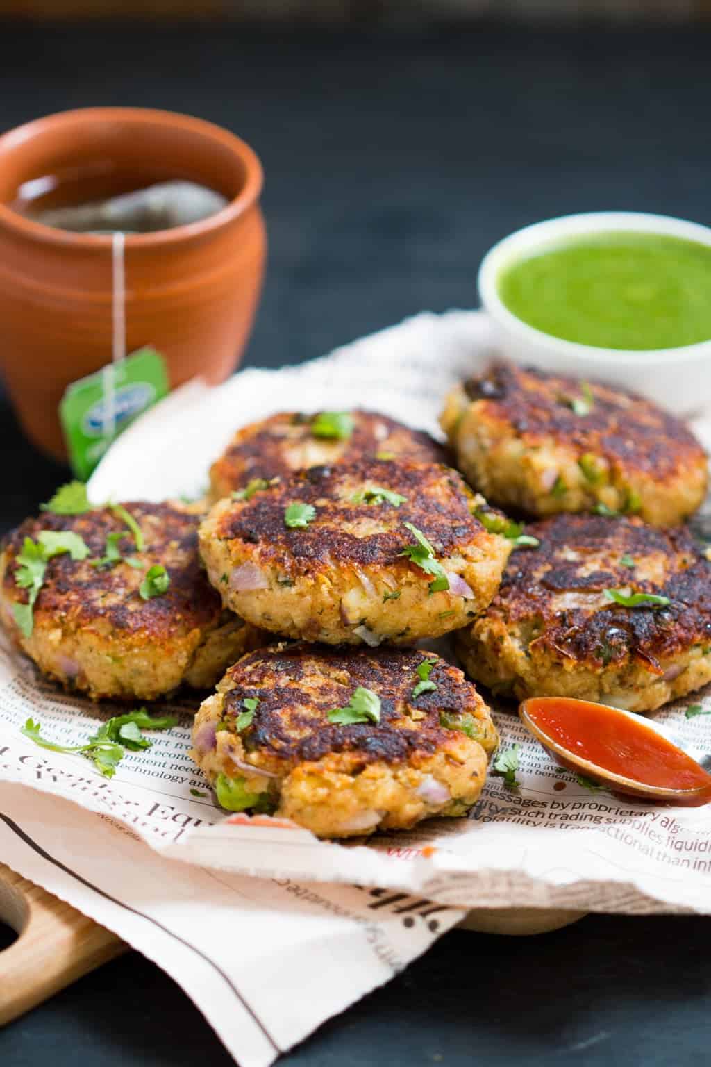 aloo paneer tikkis stacked on a brown board with newspaper alongside a small of chutney and some tea